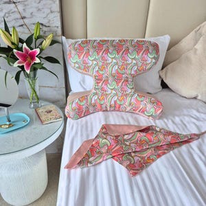 Pode incluir: Uma almofada em forma de H e um lenço triangular a condizer, ambos com um padrão abstrato multicolorido vibrante em tons de rosa, verde, laranja e branco, pousados sobre um lençol branco.
