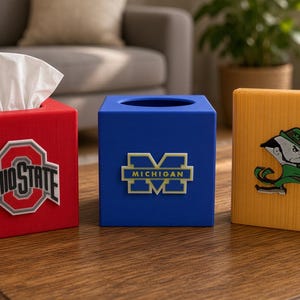 May include: Three square tissue box covers in red, blue, and yellow. The red cover features the Ohio State logo, the blue cover displays the Michigan logo, and the yellow cover shows a leprechaun mascot logo. Each box has a top opening.