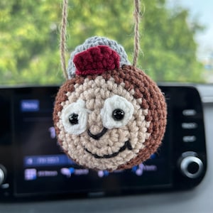May include: A crocheted ornament shaped like a smiling face, featuring a brown body, white eyes, and a grey and maroon hat. The ornament hangs from a natural twine string, ideal for car or room decoration.
