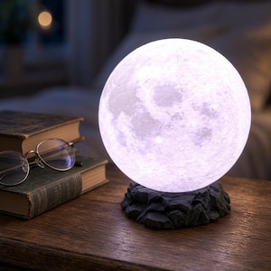 May include: A spherical moon lamp with a textured, cratered surface emitting a soft white glow. The lamp rests on a dark, jagged, rock-like base. Beside the lamp are two stacked vintage books and a pair of metal-rimmed reading glasses.