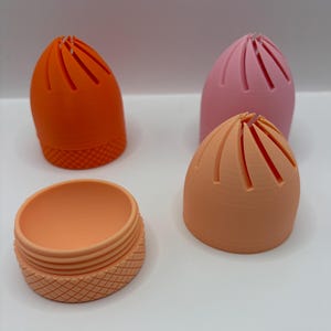 May include: Three dome-shaped containers with vertical slits at the top and a textured diamond pattern on the base. One container is orange, one is light pink, and one is peach. One peach base is separated from its matching dome-shaped lid.