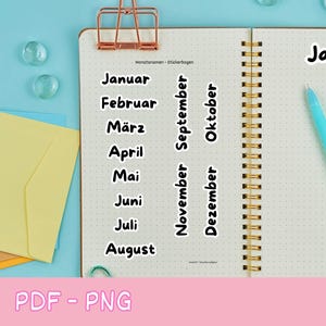 Könnte beinhalten: Ein Notizbuch mit deutschen Monatsaufklebern. Die Aufkleber enthalten die Monate Januar bis Dezember. Ein rosa herzförmiger Aufkleber sagt "Printable Planner Stickers". Ein blauer Stift und ein lila Stift sind ebenfalls sichtbar.