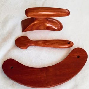 May include: Four wooden massage tools in varying shapes. The tools are made of reddish-brown wood and include a curved, crescent-shaped piece, a spoon-shaped tool, and two other pieces with handles. The tools appear to be smooth and well-crafted.