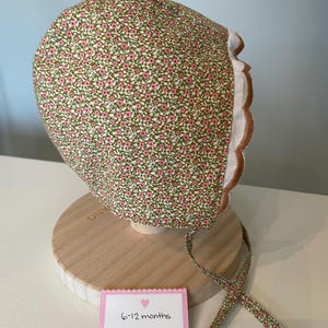 May include: A green and pink floral baby bonnet with a brown scalloped edge and white ties. The bonnet is on a wooden stand with a pink tag that says "6-12 months sweetbeebonnets".