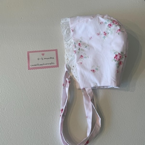 May include: A pink baby bonnet with a white lace trim and a floral pattern. The bonnet has two ties that can be tied under the chin. The bonnet is labeled "0-3 months sweetbeebonnets".
