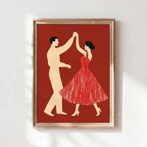 Puede incluir: Una lámina de arte enmarcada que presenta una ilustración estilizada de dos figuras bailando sobre un fondo rojo intenso sólido. Una figura aparece en color crema claro, mientras que la otra lleva un vestido rojo texturizado con detalles en blanco.