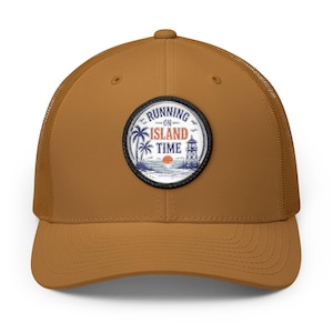 May include: A tan trucker hat featuring a circular embroidered patch on the front. The patch displays a beach scene with a palm tree, a lighthouse, and a setting sun, with the text RUNNING ON ISLAND TIME in blue and orange lettering.