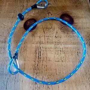 May include: A blue and yellow rope dog lead with a loop at each end and a black stopper. The lead has a metal clip and is lying on a wooden surface. The rope is a bright blue colour with yellow and green accents.