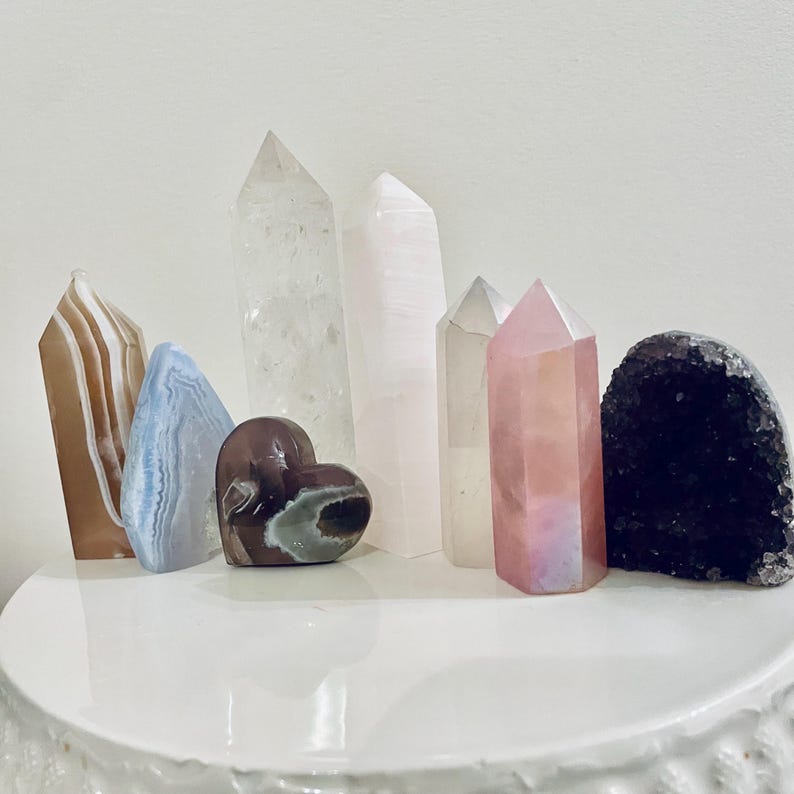 May include: A collection of various crystals and gemstones, including quartz points, a heart-shaped stone, and a geode. The crystals display a range of colors, such as clear, pink, brown, blue, and purple. They are arranged on a white surface.
