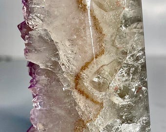 May include: A polished crystal point featuring a clear, translucent structure with internal fractures and a distinct, jagged brown mineral inclusion running vertically through the center, accented by deep purple amethyst tones along the side.