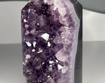 May include: A large amethyst crystal geode with a polished black side. The crystal features a deep purple color with many sparkling crystal points. The geode is a natural, decorative item.