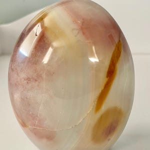 Puede incluir: Una piedra pulida de forma ovalada con una superficie lisa y brillante. La piedra presenta una mezcla de colores, incluyendo crema, rosa pálido y vetas de naranja y marrón. El aspecto general es natural y orgánico, con una sutil grieta visible.
