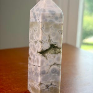 May include: A polished stone tower with a hexagonal prism shape, featuring a pattern of circular white and light gray orbs. The stone has a smooth, glossy finish and includes a small, horizontal cavity filled with sparkling, metallic gold crystals.