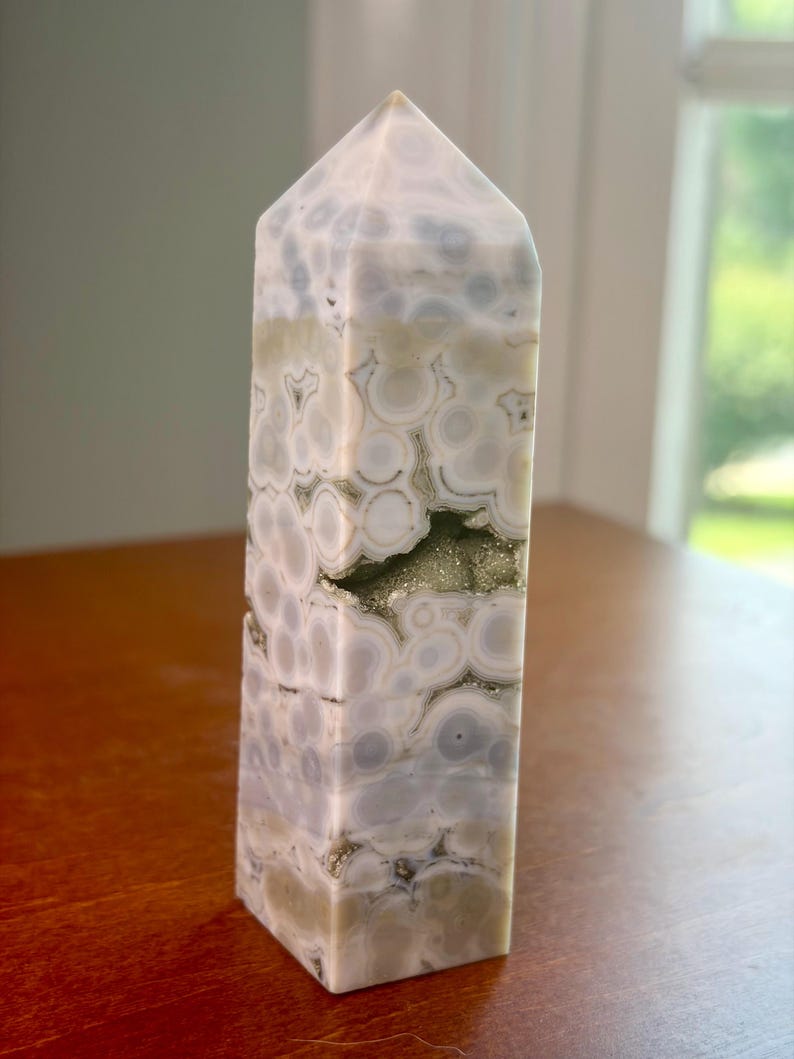 May include: A polished stone tower with a hexagonal prism shape, featuring a pattern of circular white and light gray orbs. The stone has a smooth, glossy finish and includes a small, horizontal cavity filled with sparkling, metallic gold crystals.