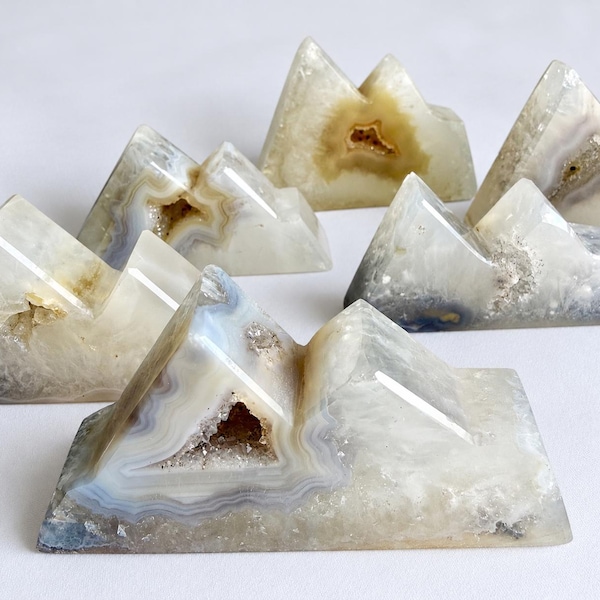 May include: A collection of six mountain-shaped sculptures carved from translucent agate stone. Each piece features natural banding in shades of white, cream, and gray, with exposed crystalline geode centers and smooth, polished surfaces.