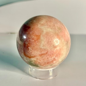 May include: A polished sphere of pink and cream-colored stone, possibly a decorative crystal ball. The orb has a smooth surface with natural variations in color and is displayed on a clear, round stand.