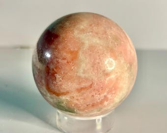 May include: A polished sphere of pink and cream-colored stone, possibly a decorative crystal ball. The orb has a smooth surface with natural variations in color and is displayed on a clear, round stand.
