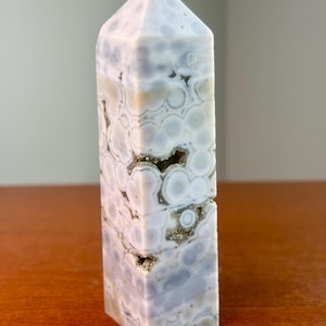 May include: A polished stone tower with a hexagonal base and a pointed top. The stone features a light gray and white orbicular pattern with circular inclusions and small, recessed crystalline cavities containing sparkling metallic mineral deposits.