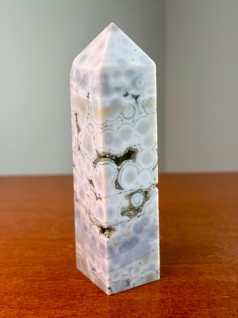 May include: A polished stone tower with a hexagonal base and a pointed top. The stone features a light gray and white orbicular pattern with circular inclusions and small, recessed crystalline cavities containing sparkling metallic mineral deposits.