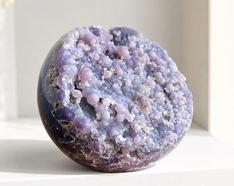 May include: A spherical mineral specimen featuring a cluster of small, rounded, light purple and lavender botryoidal crystals on its front face. The outer surface of the sphere is smooth and dark, contrasting with the textured, crystalline interior.