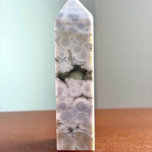 May include: A polished stone tower with a hexagonal point, featuring a pattern of circular, orb-like inclusions in shades of white, gray, and pale green. A horizontal, jagged crevice runs through the center, revealing a sparkling, dark mineral druzy.