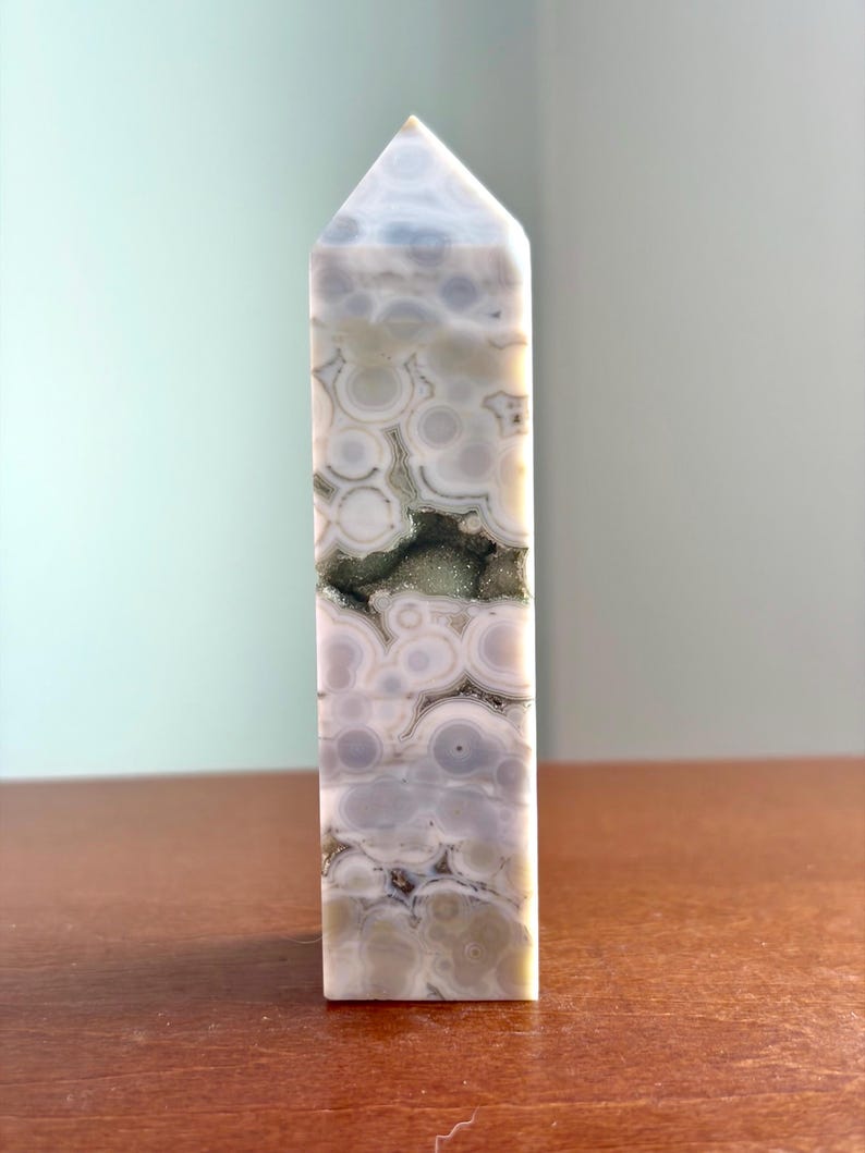 May include: A polished stone tower with a hexagonal point, featuring a pattern of circular, orb-like inclusions in shades of white, gray, and pale green. A horizontal, jagged crevice runs through the center, revealing a sparkling, dark mineral druzy.