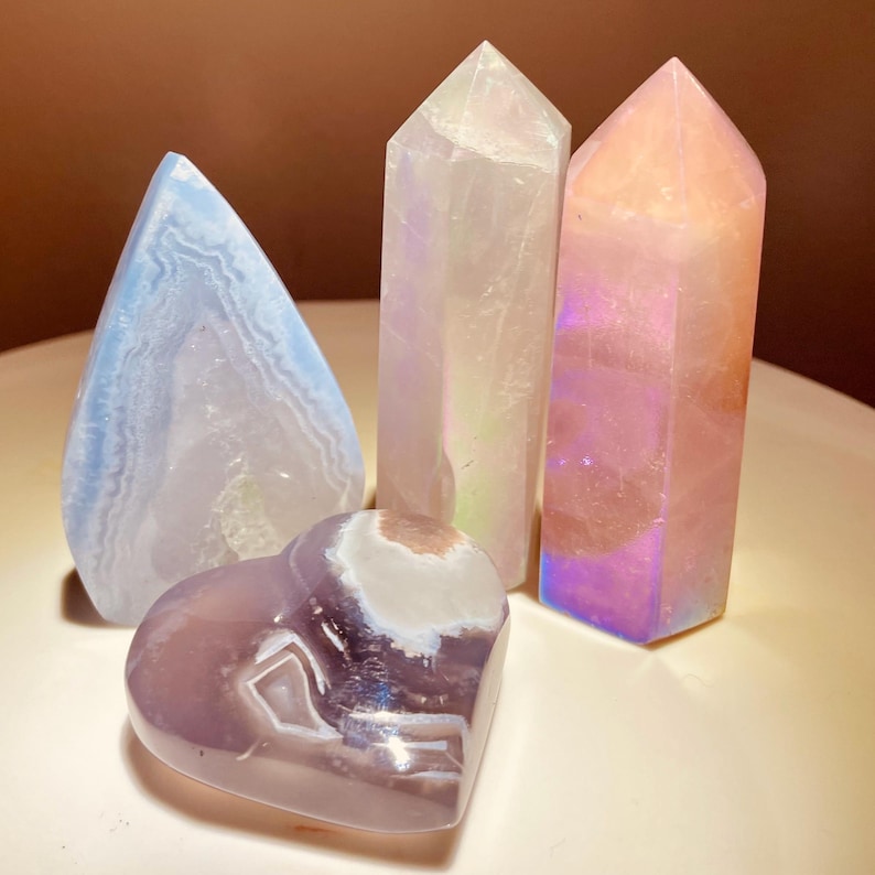 May include: Four polished crystals in shades of blue, purple, and pink. The collection includes a heart-shaped stone, a teardrop-shaped stone, and two tower-shaped crystals. The crystals have a translucent quality and are arranged on a white surface.
