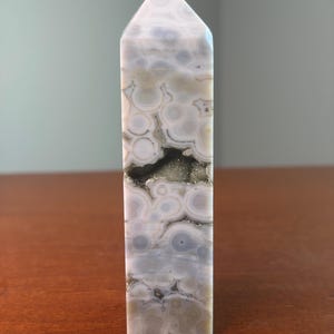 May include: A polished stone tower with a hexagonal point, featuring a cream and light gray orbicular pattern with concentric circles. A horizontal, jagged cavity runs through the center, revealing a sparkling, metallic gold-toned druzy interior.