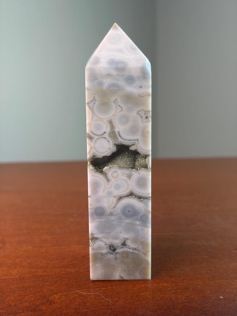 May include: A polished stone tower with a hexagonal point, featuring a cream and light gray orbicular pattern with concentric circles. A horizontal, jagged cavity runs through the center, revealing a sparkling, metallic gold-toned druzy interior.