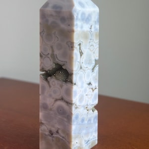May include: A polished stone tower with a hexagonal point, featuring a light gray and lavender color palette with circular orbicular patterns and a central horizontal crevice containing small, sparkling, dark gray crystalline mineral deposits.