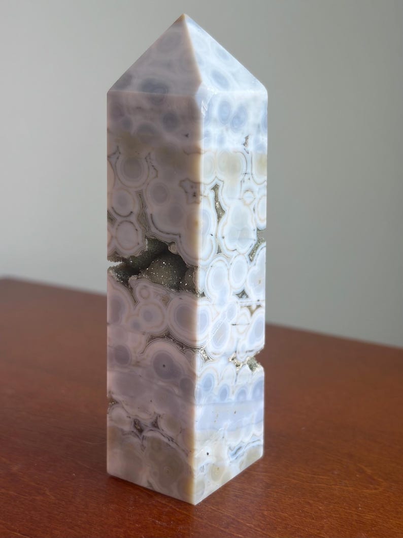 May include: A polished stone tower with a hexagonal point, featuring a light gray and lavender color palette with circular orbicular patterns and a central horizontal crevice containing small, sparkling, dark gray crystalline mineral deposits.