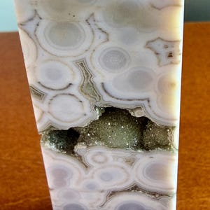 May include: A polished stone tower featuring a light gray and white orbicular pattern with concentric circular bands. A horizontal fissure runs across the center, revealing a textured, sparkling interior cavity filled with small green crystals.