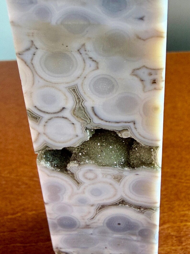 May include: A polished stone tower featuring a light gray and white orbicular pattern with concentric circular bands. A horizontal fissure runs across the center, revealing a textured, sparkling interior cavity filled with small green crystals.