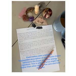 May include: Assortment of crystals and gemstones, including Mookaite Jasper spheres, Garden Quartz, and Crazy Lace Agate carvings. A handwritten note describes each stone's energy. A pen and a mug are also visible.