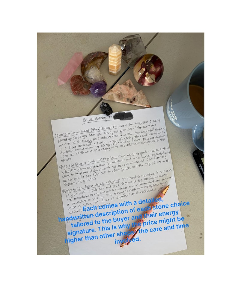 May include: Assortment of crystals and gemstones, including Mookaite Jasper spheres, Garden Quartz, and Crazy Lace Agate carvings. A handwritten note describes each stone's energy. A pen and a mug are also visible.