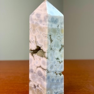 May include: A polished stone tower with a hexagonal point, featuring a pale lavender and white orbicular pattern. The surface displays circular inclusions and a central horizontal crevice filled with small, sparkling, light-colored mineral crystals.