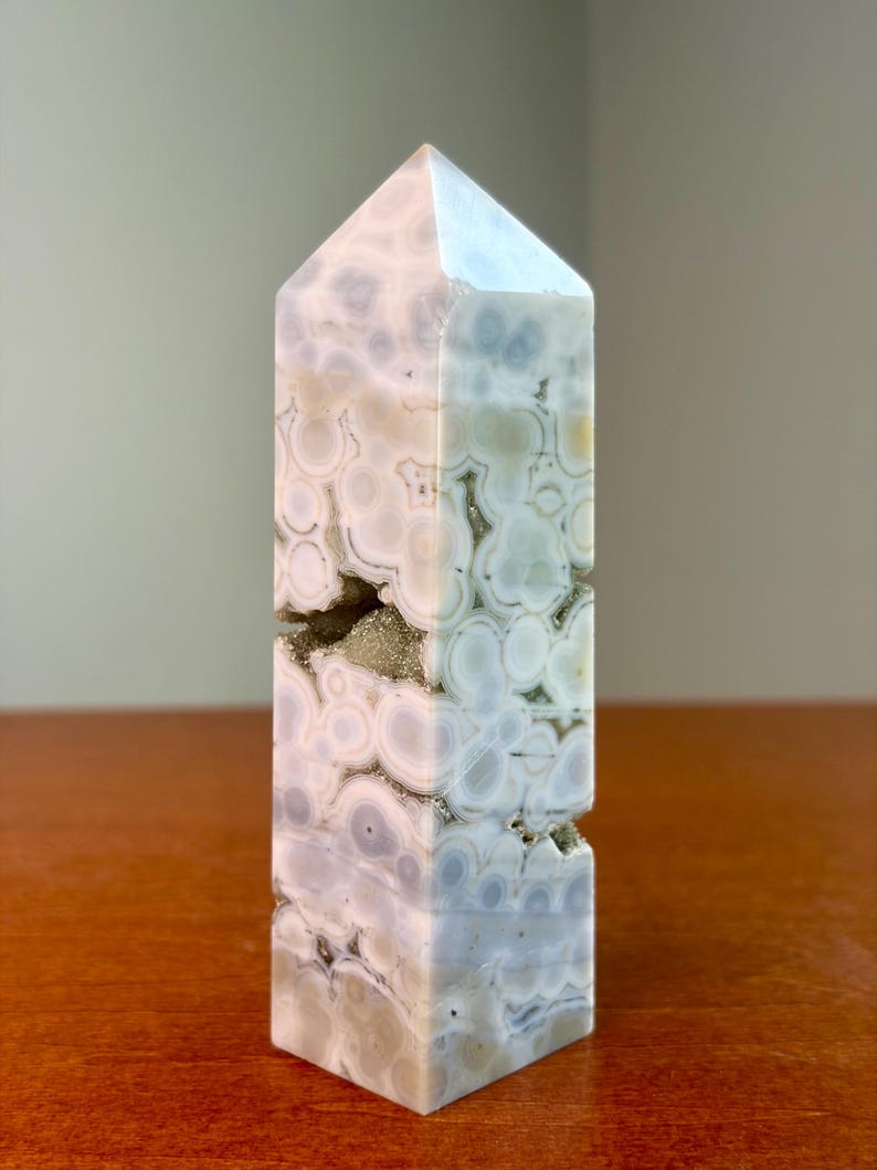 May include: A polished stone tower with a hexagonal point, featuring a pale lavender and white orbicular pattern. The surface displays circular inclusions and a central horizontal crevice filled with small, sparkling, light-colored mineral crystals.