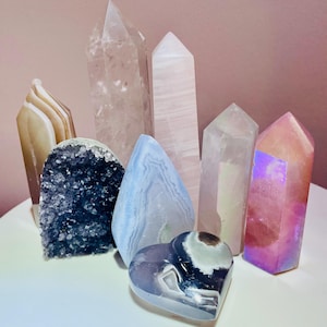 May include: A collection of various crystals and gemstones, including quartz points, a heart-shaped stone, and a geode. The crystals are in shades of pink, clear, blue, and iridescent colors. The arrangement is on a white surface against a pink background.