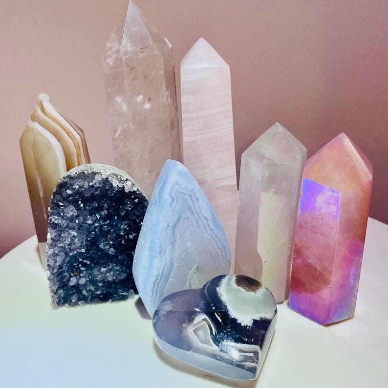 May include: A collection of various crystals and gemstones, including quartz points, a heart-shaped stone, and a geode. The crystals are in shades of pink, clear, blue, and iridescent colors. The arrangement is on a white surface against a pink background.