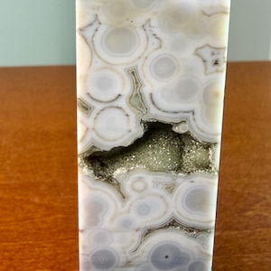 May include: A polished stone tower featuring a natural pattern of concentric circles in shades of white, cream, and soft gray. A horizontal, jagged crevice runs through the center, revealing a cluster of small, sparkling, light green crystals.