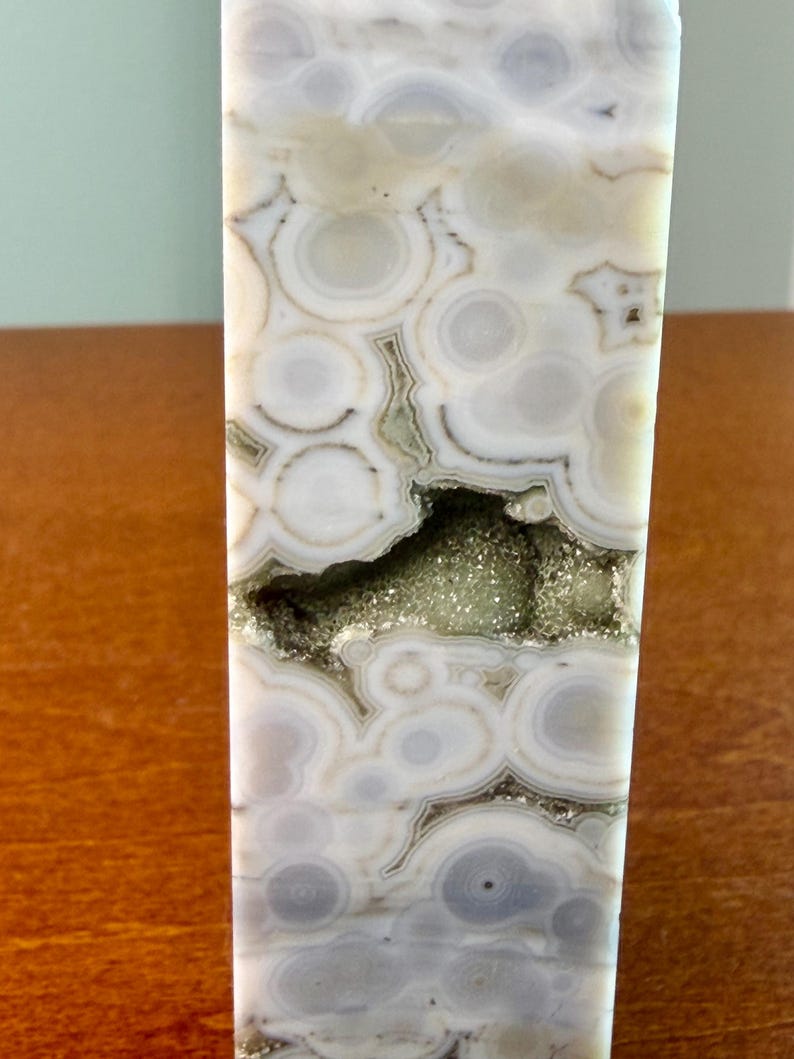May include: A polished stone tower featuring a natural pattern of concentric circles in shades of white, cream, and soft gray. A horizontal, jagged crevice runs through the center, revealing a cluster of small, sparkling, light green crystals.