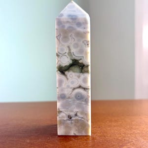 May include: A polished stone tower with a pointed top, featuring a light gray and white orbicular pattern with concentric circles. A horizontal, jagged crevice runs through the center, revealing a dark, textured, crystalline mineral interior.