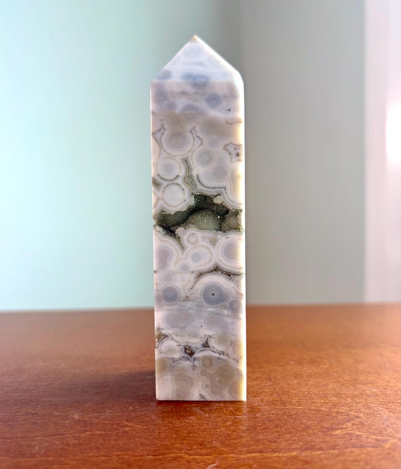May include: A polished stone tower with a pointed top, featuring a light gray and white orbicular pattern with concentric circles. A horizontal, jagged crevice runs through the center, revealing a dark, textured, crystalline mineral interior.