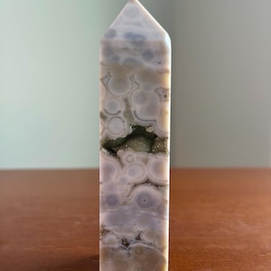 May include: A polished stone tower with a pointed top, featuring a light gray and cream color palette with circular orbicular patterns throughout. A horizontal, jagged crevice runs through the center, revealing a sparkling, dark crystalline interior.