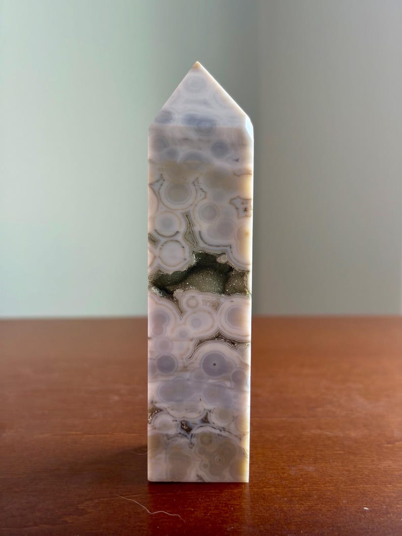 May include: A polished stone tower with a pointed top, featuring a light gray and cream color palette with circular orbicular patterns throughout. A horizontal, jagged crevice runs through the center, revealing a sparkling, dark crystalline interior.