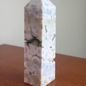 May include: A polished stone tower with a pointed top, featuring a light lavender and cream-colored pattern of circular orbs. A horizontal fissure runs through the center of the stone, revealing a textured, sparkling crystalline interior.
