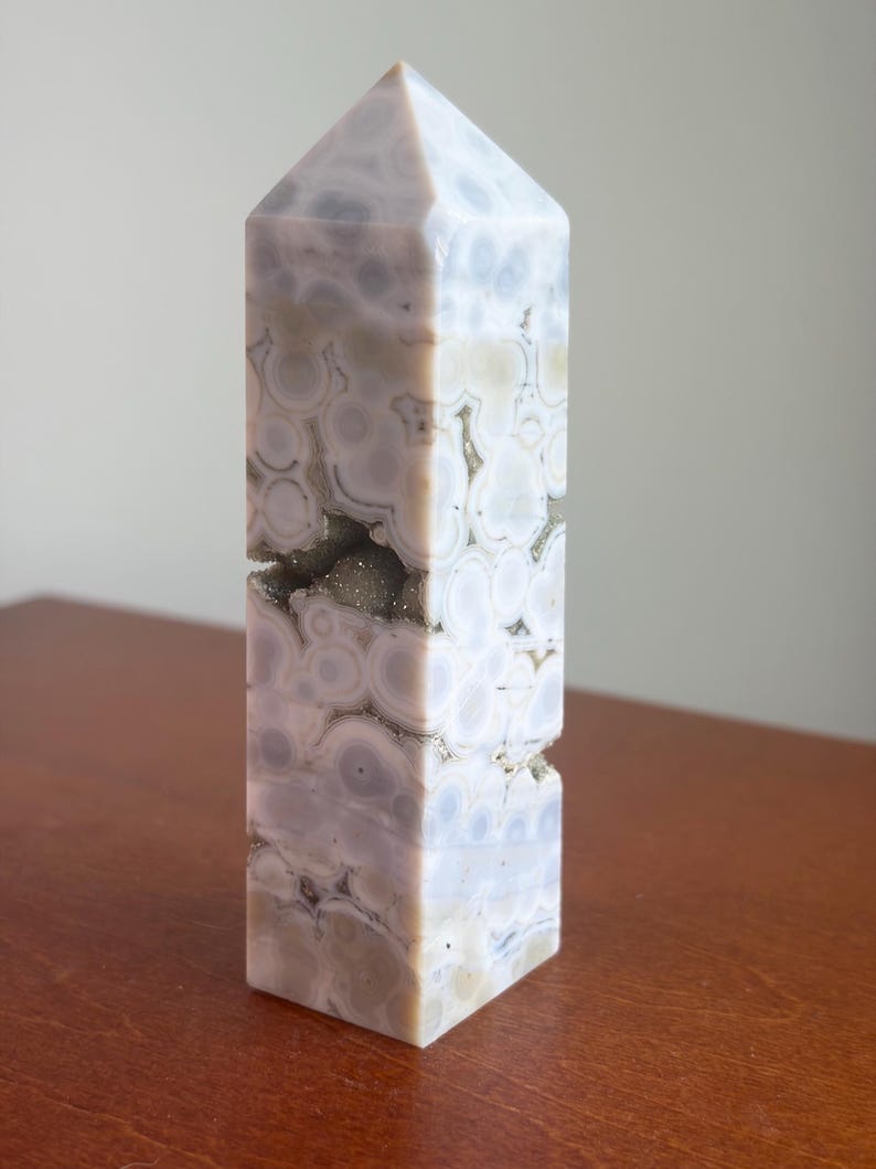 May include: A polished stone tower with a pointed top, featuring a light lavender and cream-colored pattern of circular orbs. A horizontal fissure runs through the center of the stone, revealing a textured, sparkling crystalline interior.