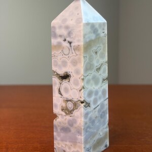 May include: A polished stone tower with a hexagonal point, featuring a complex pattern of circular, orb-like inclusions in shades of pale lavender, cream, and soft gray. The surface displays natural crystalline cavities with metallic flecks.