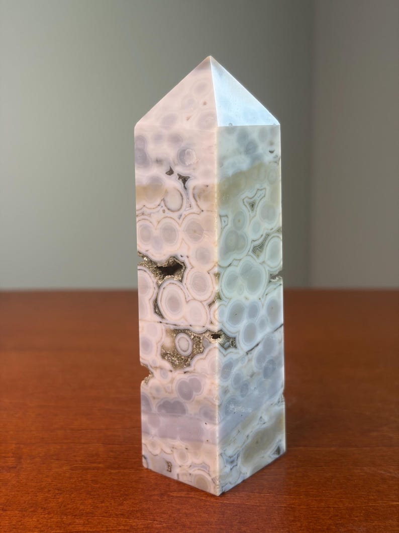 May include: A polished stone tower with a hexagonal point, featuring a complex pattern of circular, orb-like inclusions in shades of pale lavender, cream, and soft gray. The surface displays natural crystalline cavities with metallic flecks.