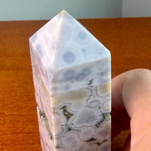 May include: A polished stone tower with a pointed top, featuring a natural pattern of circular, orb-like inclusions in shades of pale blue, white, and soft beige, with small crystalline crevices visible along the vertical sides of the mineral.